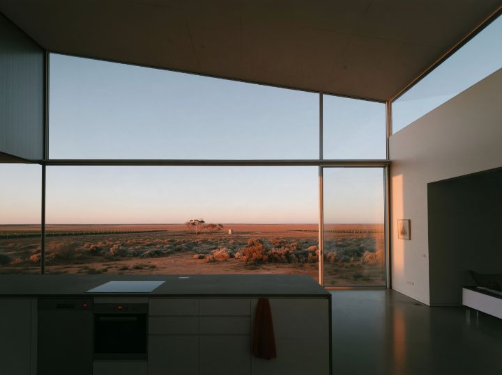 A pale kitchen looking over saltbush and red earth to the vines, the roof rising towards the glass